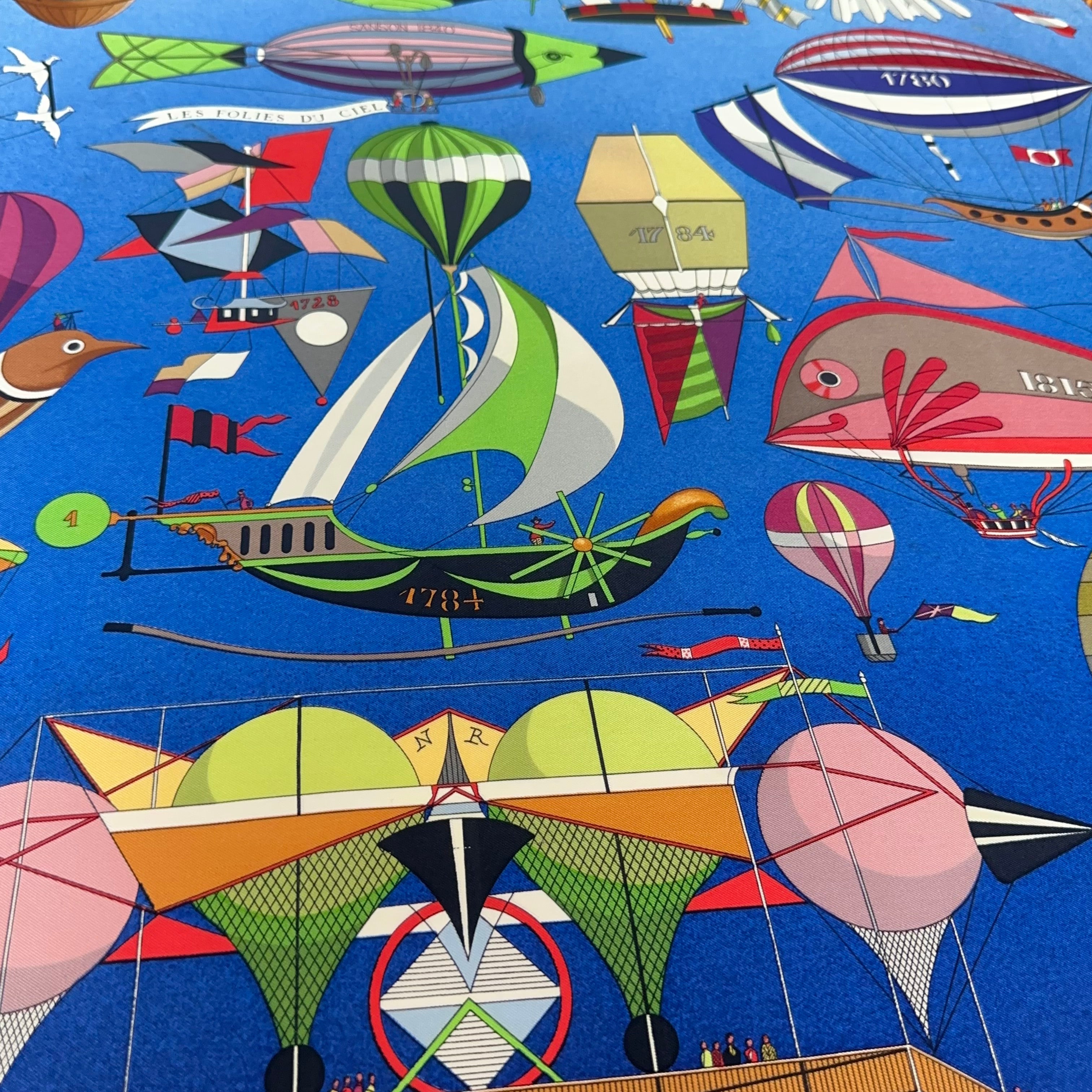 Vue centrale du carré illustrant l'histoire de l'aviation : navire aérien à voiles vertes (1784), dirigeable poisson rose (1815) et ballon complexe sur fond bleu.