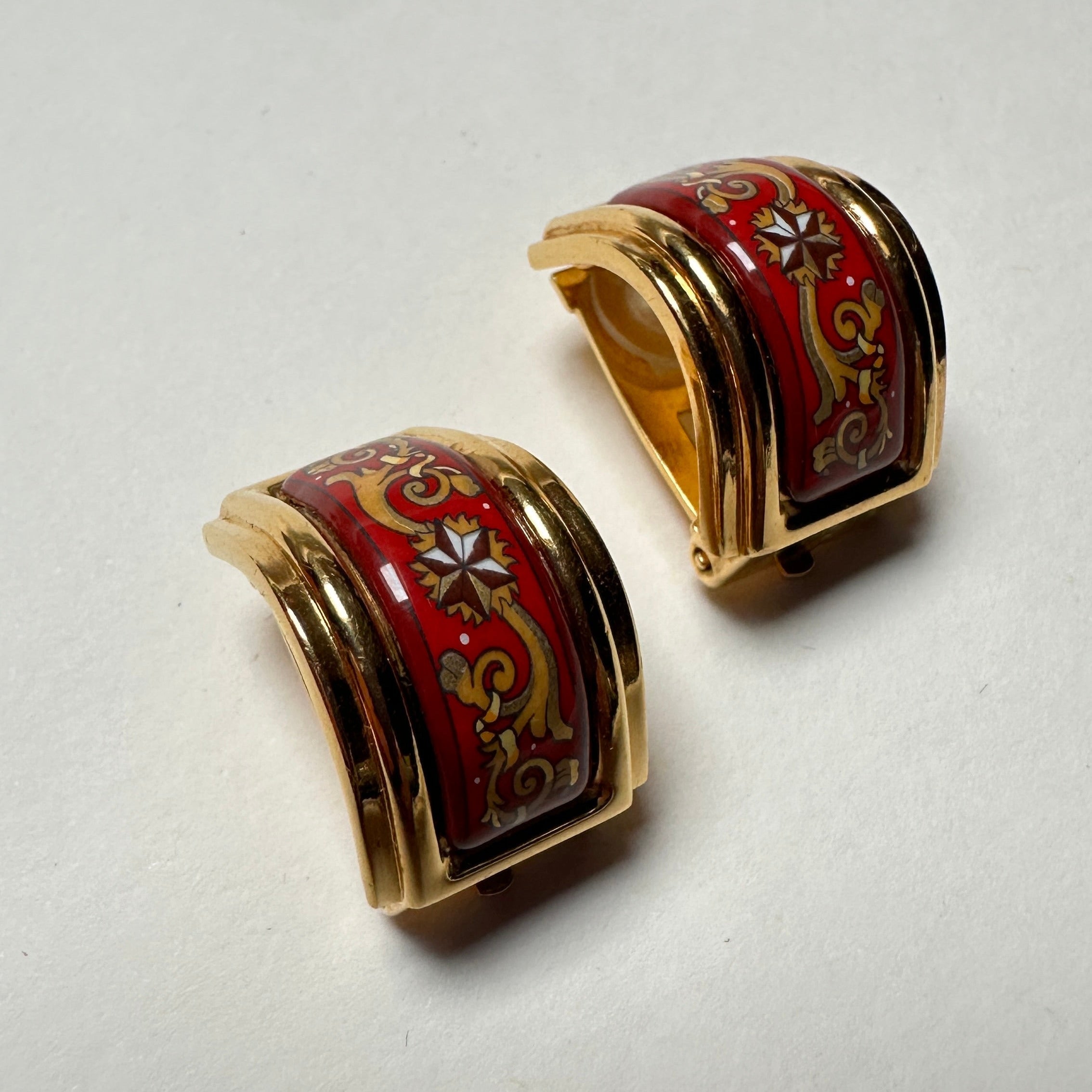 Gros plan détaillé montrant la qualité de l'émail grand feu rouge, les volutes dorées et le motif étoile central sur les boucles d'oreille clip Hermès.