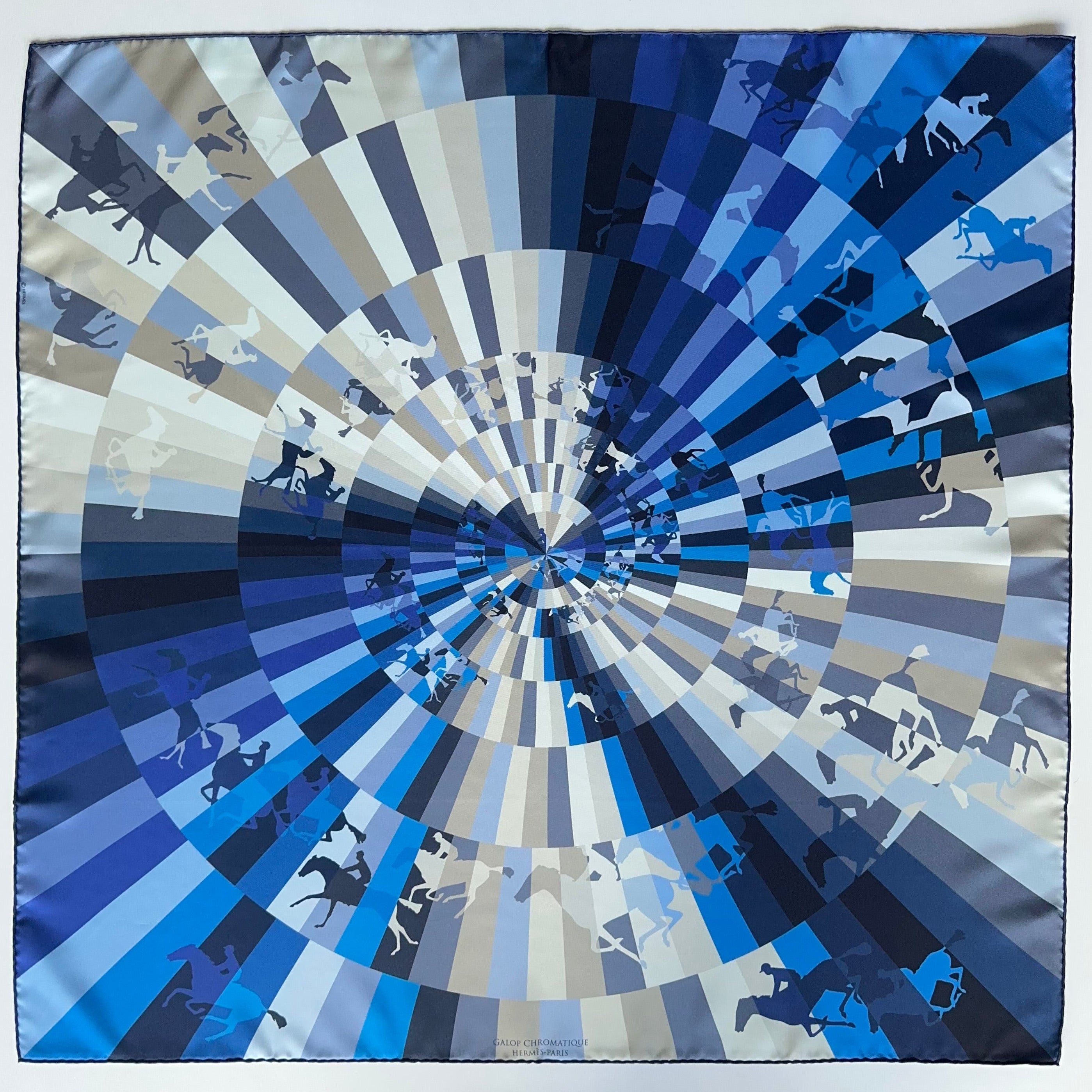 Vue d'ensemble du Carré Hermès "Galop Chromatique" sur fond blanc. Motif cinétique circulaire représentant des chevaux au galop en dégradé de bleus et gris.